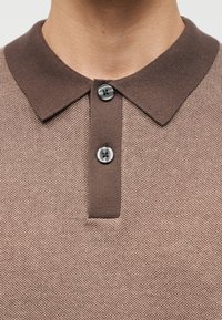 Κοντινό πλάνο ενός ατόμου που φοράει καφέ πλεκτό πόλο πουκάμισο με υφή, ριμπ γιακά και δύο μαύρα κουμπιά κουμπωμένα στον λαιμό.