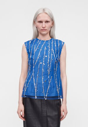 Femme aux cheveux pâles portant un haut bleu sans manches avec des rayures verticales en sequins argentés et une jupe noire, debout devant un fond blanc.
