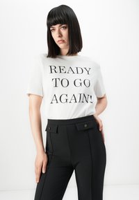Young woman with straight dark hair wearing white shirt with "READY TO GO AGAIN!" text and high-waisted black pants with buttoned pockets.