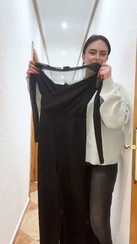 Γυναίκα με λευκό πουλόβερ κρατάει ένα μαύρο αμάνικο φόρεμα με διακριτική λεπτομέρεια από διχτυωτό ύφασμα στο πάνω μέρος, στέκεται σε διάδρομο με λευκούς τοίχους και πλακιδωτό δάπεδο.