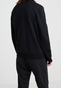 Sudadera negra con cuello redondo, puños y dobladillo acanalados, y una textura suave; diseño minimalista sin patrones o logotipos visibles.