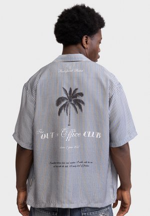 Homme portant une chemise à manches courtes rayée bleu et blanc avec un palmier et un texte blanc imprimés dans le dos, tourné dos à la caméra.
