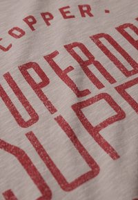 Tela de color gris claro con texto rojo impreso prominente. El texto dice "SUPERB" con un acabado desgastado, lo que sugiere una apariencia casual y texturada.