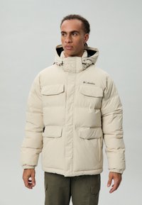 Beige pufferjack met een hoge kraag, grote voorzakken en een trekkoordcapuchon. Gemaakt van een gladde, geïsoleerde stof voor warmte.