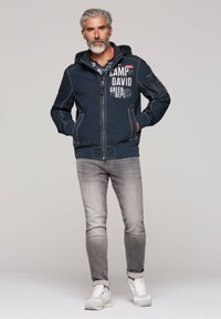 Navy zip-up jacket with hood, featuring white stitching and printed text; paired with light gray jeans and white sneakers.