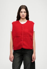 Sarkans fleeces vestes ar apaļu kaklu un priekšējiem pogām, ar diviem lieliem kabatām un mīkstu, teksturētu virsmu. Valkāta virs baltas kreklas.