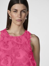 Lys rosa ärmlös topp med texturerad, volangigt tyg i blommönster. Modellen har långt hår och silvriga hoopörhängen.