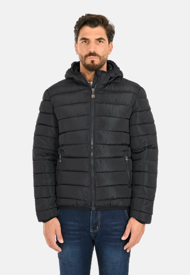 Uomo che indossa una giacca nera trapuntata con zip frontale e cappuccio, abbinata a jeans blu scuro, in piedi contro uno sfondo chiaro e uniforme.