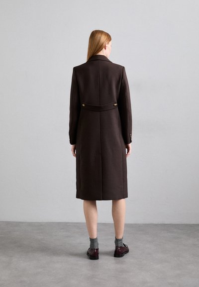 Manteau en laine marron avec une silhouette ajustée, doté d'une ceinture au dos et de boutons dorés, descendant jusqu'au mollet, porté sur des chaussettes grises et des chaussures.