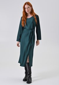 Abito di satin verde scuro con maniche lunghe, dettaglio a nodo in vita e spacco laterale, abbinato a stivali neri fino al ginocchio.