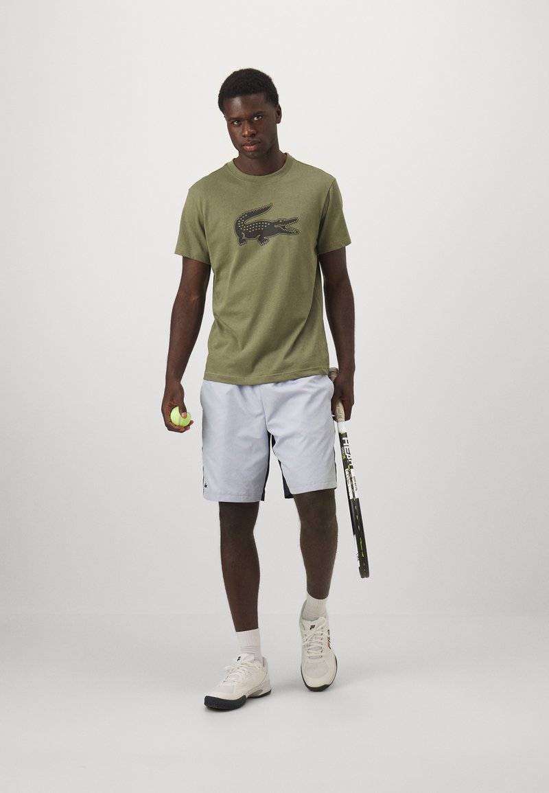 Olijfgroenen katoenen t-shirt met zwart krokodillenlogo, gecombineerd met lichtgrijze shorts, witte tennisschoenen, houdend een tennisracket en een bal.