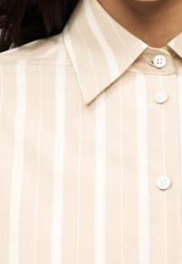 Gros plan d'une chemise rayée verticalement beige et blanche avec col pointu et trois boutons blancs visibles.