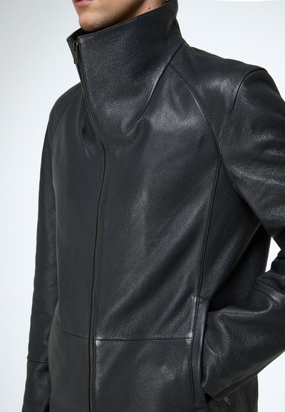 Mann trägt eine schwarze Lederjacke mit hohem Reißverschlusskragen, die hochgeschlossen ist, und hat die Hände in den Jackentaschen.