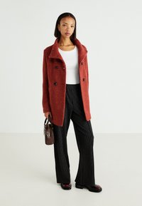 Red wool-blend double-breasted coat with black buttons, featuring a wide collar. Paired with black-striped trousers and patent leather ankle boots.