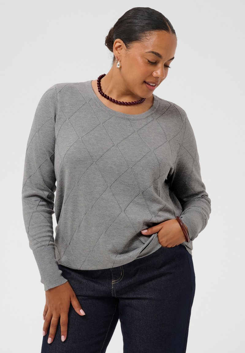 Femme portant un pull gris à motif losanges et un jean foncé, avec un collier et un bracelet de perles bordeaux, posant avec une main dans la poche.