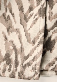Beige tyg med kamouflage-mönster i nyanser av brunt och grått. Mjuk textur, detaljerad söm synlig längs kanten.