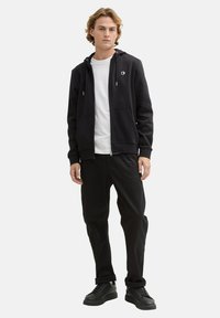 Felpa nera con zip e coulisse, con texture a coste, e tasche frontali, indossata sopra una t-shirt bianca e abbinata a pantaloni neri e sneakers.