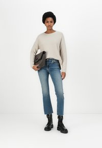 Suéter de malha bege, jeans de cintura alta azuis, botas de combate pretas e uma carteira escura. O suéter tem um decote redondo e um corte descontraído.