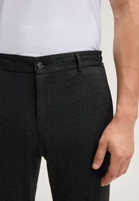Pantaloni grigio scuro con tessuto testurizzato, vita elastica e chiusura con bottone. La mano è appoggiata sul tessuto per dare un'idea delle dimensioni.