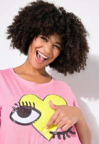 Camiseta de algodón rosa con un diseño de corazón amarillo y un ojo, con contornos y pestañas negras. La tela es suave y tiene un corte holgado.