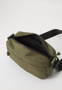 Olive green crossbody bag with a zip closure, featuring a smooth texture, adjustable strap, and logo label inside.