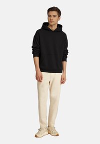 Mustat hupparit, joissa on etutasku, on yhdistetty vaaleisiin beigen värisiin housuihin ja vaaleisiin lenkkareihin. Malli seisoo yksinkertaisella taustalla.