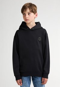 Haut à capuche noir avec une grande capuche, des poignets et un ourlet côtelés, arborant un petit logo circulaire sur la poitrine, associé à un jean bleu clair.