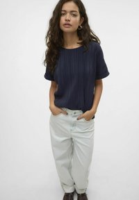 Marineblå ribbet t-shirt med korteærmer, parret med lyseblå højtaljede jeans. Modellen står med hænderne i lommerne iført brune støvler.