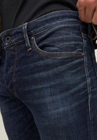 Krupni plan tamnoplave traperice s vidljivim prednjim džepom, detaljima šivanja i rukom koja podešava pojas.