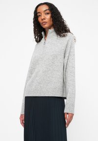 Suéter gris con cremallera en el cuello, de manga larga y con dobladillo acanalado, llevado sobre una falda oscura plisada. Textura suave con un ajuste relajado.