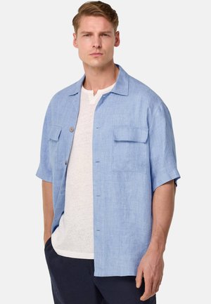 Helles blaues Kurzarmhemd aus strukturiertem Stoff, mit zwei Brusttaschen und einem Button-Down-Kragen, getragen über einem weißen Unterhemd.