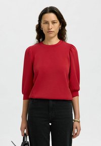 Blusa rossa a maniche corte con scollatura rotonda e maniche a palloncino. Indossata con jeans neri e porta una borsa nera. Tessuto dalla consistenza liscia.