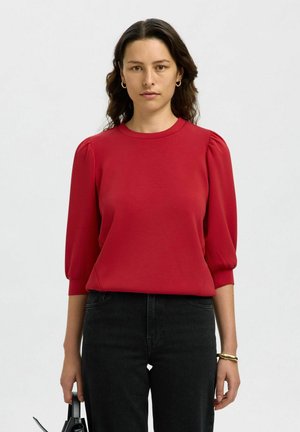 Red, short-sleeve blouse with a round neckline and puffed sleeves. Worn with black jeans and holding a black bag. Smooth fabric texture.
