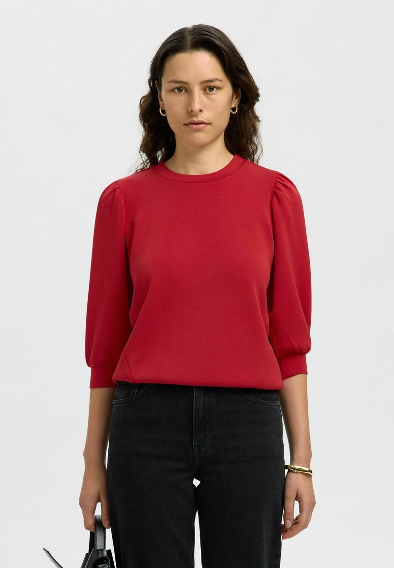 Blusa rossa a maniche corte con scollatura rotonda e maniche a palloncino. Indossata con jeans neri e porta una borsa nera. Tessuto dalla consistenza liscia.