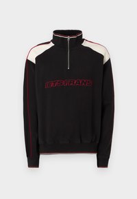 Μαύρη μπλούζα φόρμας με ψηλό γιακά και φερμουάρ τετάρτου. Διαθέτει κόκκινα και κρέμ χρώματα σε μπλοκ και κείμενο "LETS FRANS" σε έντονο κόκκινο. Λεπτομέρειες σε κόκκινο χρώμα.