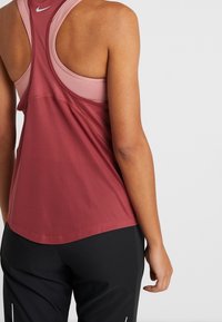 Różowy top sportowy o kolorze malinowym z siateczkową teksturą, z tyłem typu racerback i kontrastującymi jaśniejszymi ramiączkami; w parze z czarnymi spodniami z białymi akcentami.