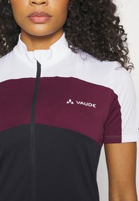 Maillot de cyclisme à manches courtes en blanc, bordeaux et noir. Doté d'un col haut et d'une fermeture éclair. Logo "Vaude" affiché à l'avant.