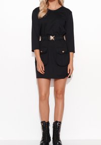 Vestido negro con mangas tres cuartos, una cintura ceñida con un cinturón de hebilla dorada y dos grandes bolsillos. Combinado con botas negras con cordones.
