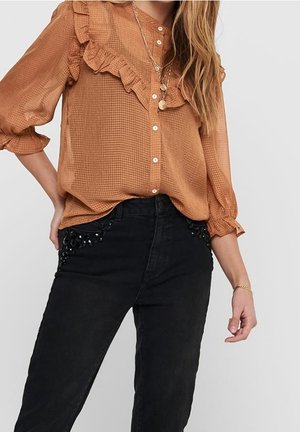 Woman wearing a brown ruffled blouse with white buttons and layered necklaces, paired with black pants featuring beaded pocket details.