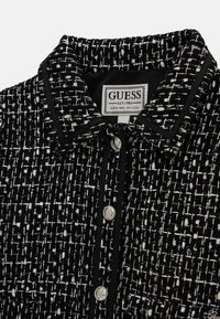 Czarno-biała tkanina tweedowa z kołnierzem, ozdobiona srebrnymi guzikami i czarną lamówką wzdłuż krawędzi. Widoczna metka z logo GUESS.