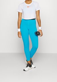 Conjunto de ropa deportiva que incluye una camiseta recortada blanca con un logo naranja de Nike y leggins turquesas. Los accesorios incluyen una muñequera y una botella de agua.