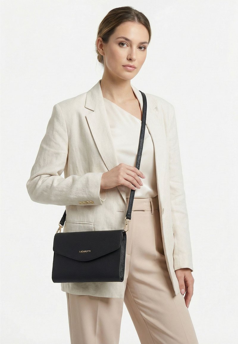 Mujer con blazer y pantalones claros, sosteniendo un bolso bandolera negro con solapa y logo dorado que dice Lazarotti.