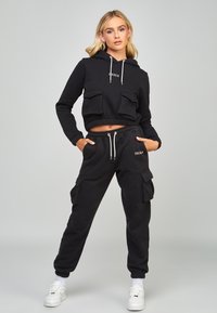 Felpa nera corta con tasche frontali e cordino, abbinata a pantaloni joggers neri coordinati con tasche cargo laterali e polsini elasticizzati.