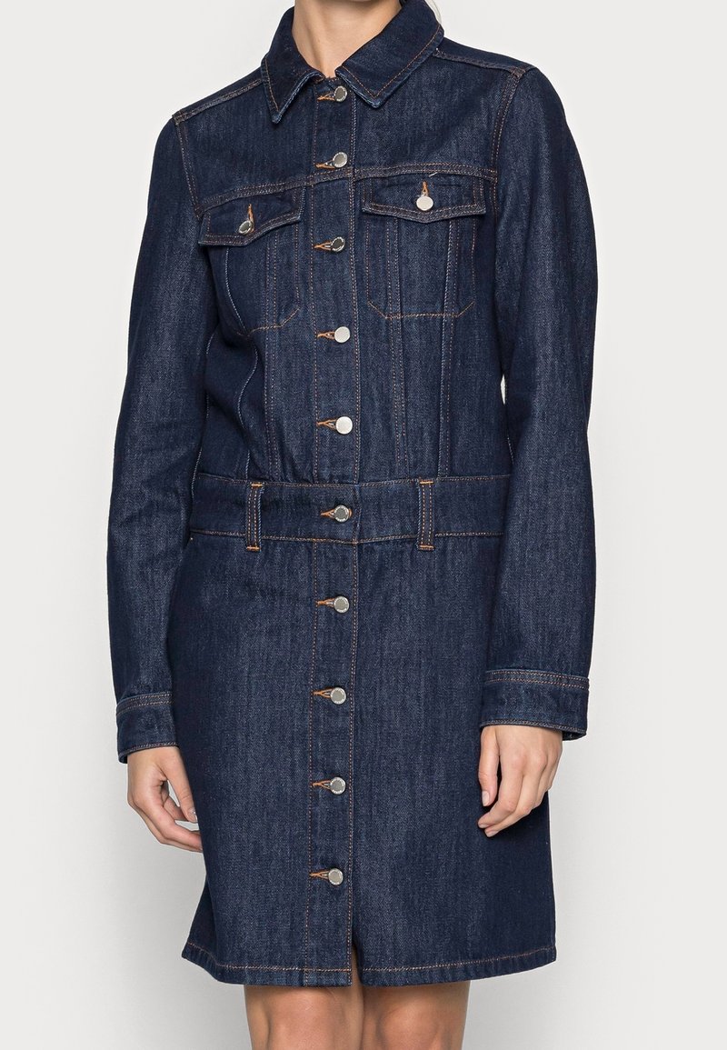 Woman wearing a dark blue denim button-up dress with long sleeves, chest pockets, and orange stitching, standing against a plain background.