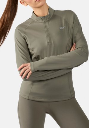 Frau trägt olivgrünes, langärmeliges Sportoberteil mit Reißverschluss und passende Leggings, verschränkte Arme, vor weißem Hintergrund stehend.