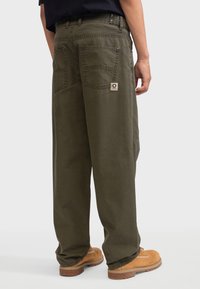 Pantalons cargo en coton vert olive avec une coupe décontractée, deux poches arrière et un petit patch de marque près de la poche droite. Portés avec des bottes marron.