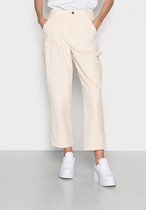 Persoon die off-white corduroy broek draagt met voor- en zijzakken, gecombineerd met witte platformsneakers en een ingestoken wit overhemd.