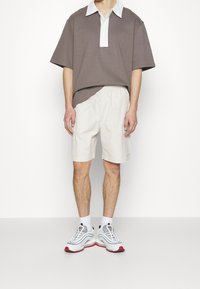 Hnědá polokošile s krátkým rukávem a bílým límečkem, spárovaná se krémovými šortkami a šedými teniskami s červenými akcenty a texturovanou podrážkou.