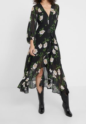 Schwarzes Blumenkleid mit langen transparenten Ärmeln, tiefem V-Ausschnitt und Rüschensaum. Enthält weiße Blumen und grüne Blätter. Kombiniert mit schwarzen Stiefeln.
