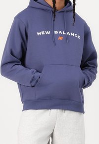 Lila hoodie med kängurufickor, har vit text "NEW BALANCE" och en liten orange logotyp. Ribbad mudd vid ärmarna och nederkant, tillverkad av mjukt tyg.